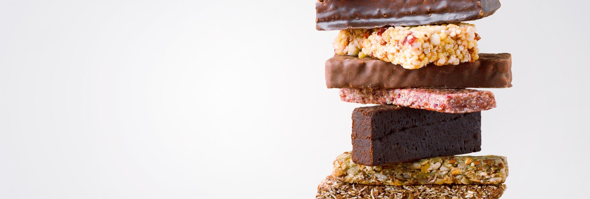 A tall stack of assorted snack and protein bars with various textures, colors, and coatings, including chocolate, oats, seeds, and fruit pieces, arranged on a light background.