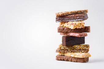 A tall stack of assorted snack and protein bars with various textures, colors, and coatings, including chocolate, oats, seeds, and fruit pieces, arranged on a light background.